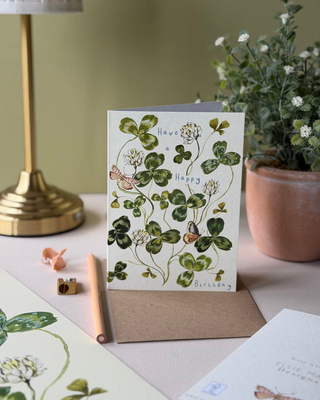 A botanical greeting card with a white background and green clover illustration, displayed on a desk with a kraft envelope, a lamp, a potted plant, and a pencil.