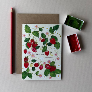 A strawberry-themed greeting card with the text 'Have the sweetest day' is displayed on a light gray surface next to a brown envelope, a red pencil, and two small paint containers.