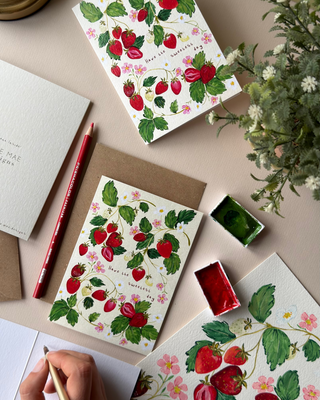 A styled flat-lay of strawberry-themed stationery, including a notebook, card, and envelope with a watercolor illustration of red strawberries and green leaves. A hand holding a gold pen is visible in the bottom-left corner, and the scene is styled with a pencil, paint palette, and greenery on a beige surface.