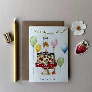 A watercolor birthday card featuring a strawberry cake with balloons and the text 'Make a wish', shown with a brown envelope on a neutral background alongside a gold pencil, a wooden sharpener, a white flower sticker, and a strawberry sticker.
