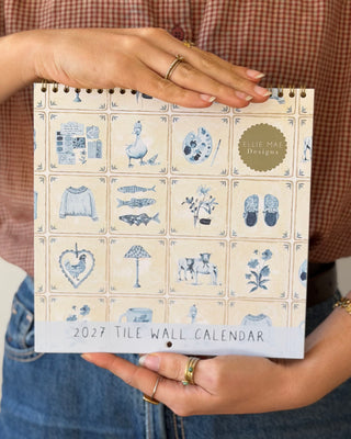 A person holds a 2027 Tile Wall Calendar featuring a vintage-inspired blue and cream tile pattern. 