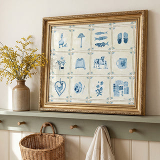 A large framed art print featuring blue and white Delft-style ceramic tiles with hand-drawn blue illustrations, displayed on a green shelf in a styled room setting.