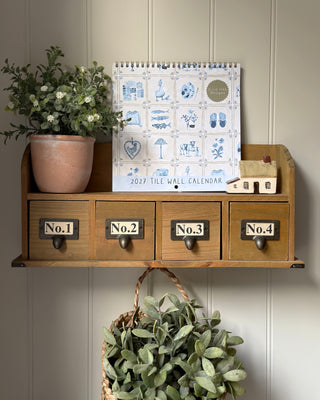 A 2027 tile wall calendar with blue illustrations stands on a wooden shelf with four labeled drawers. A potted plant sits on the left, and a small house-shaped object is on the right.