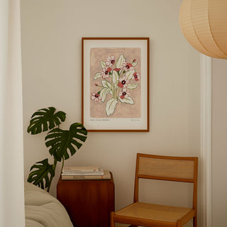 Primrose art print, a dusky light pink painted background with rosey pink and deep maroon flowers and olive green leaves. With small text reading floral studies:primrose NO.4styled in a dark wood frame in a bedroom