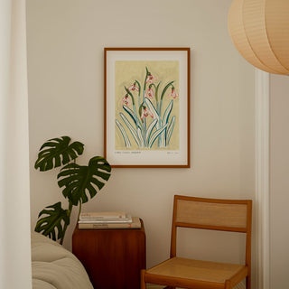 Snowdrop art print, a light muted yellow painted background with pinky snowdrop flowers and olive green/ turquoise leaves. With small text reading floral studies: snowdrop no.2 styled in a dark wood frame in a bedroom