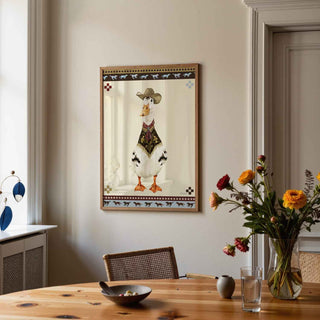 A framed art print featuring a white goose dressed in a cowboy hat and vest, hanging on a beige wall in a styled dining room with a wooden table and flowers.