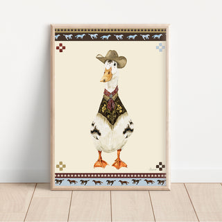 A framed art print featuring a western-style duck wearing a cowboy hat and patterned vest, leaning against a white wall on a light wood floor.