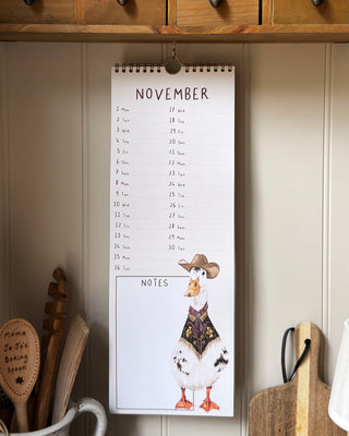 A spiral-bound November calendar page hanging on a kitchen wall, featuring a printed duck wearing a cowboy hat and vest in the notes section. Wooden cabinets and kitchen utensils are visible around it.