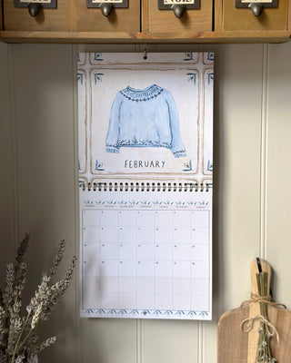 A spiral-bound wall calendar hanging on a beige paneled wall, featuring a watercolor illustration of a light blue sweater on the February page. The calendar is styled in a rustic kitchen setting with wooden cabinets, dried flowers, and a burlap cutting board.