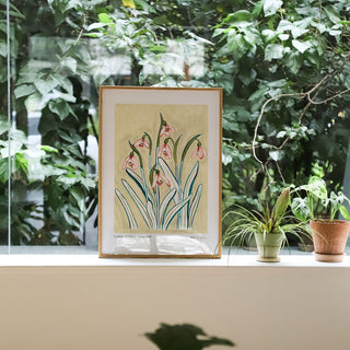 Snowdrop art print, a light muted yellow painted background with pinky snowdrop flowers and olive green/ turquoise leaves. With small text reading floral studies: snowdrop no.2 styled in a think light oak frame against greenery and plants