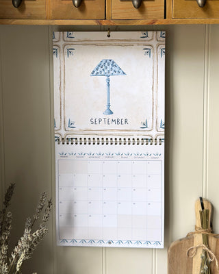 A hanging spiral-bound September desk calendar with a vintage-style illustration of a blue patterned lamp and decorative border, displayed against a cream paneled wall.