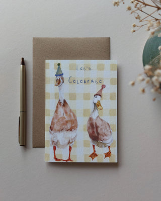 Greeting card with ducks wearing party hats on a checkered background, next to a pen and dried flowers.