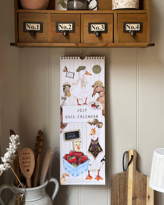 A vertical 2027 Duck Calendar hanging on a cream paneled wall in a rustic kitchen setting, featuring whimsical duck illustrations and a spiral binding at the top. Above it is a wooden cabinet with four drawers labeled No.