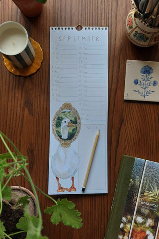 Calendar with a duck illustration on a wooden table with plants and a candle.