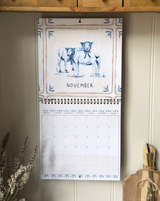A spiral-bound November desk calendar hanging on a wall, featuring a blue-and-cream illustration of two sheep and decorative border details. The calendar is photographed in a real interior setting with wood cabinetry, dried flowers, and a cutting board visible around it.