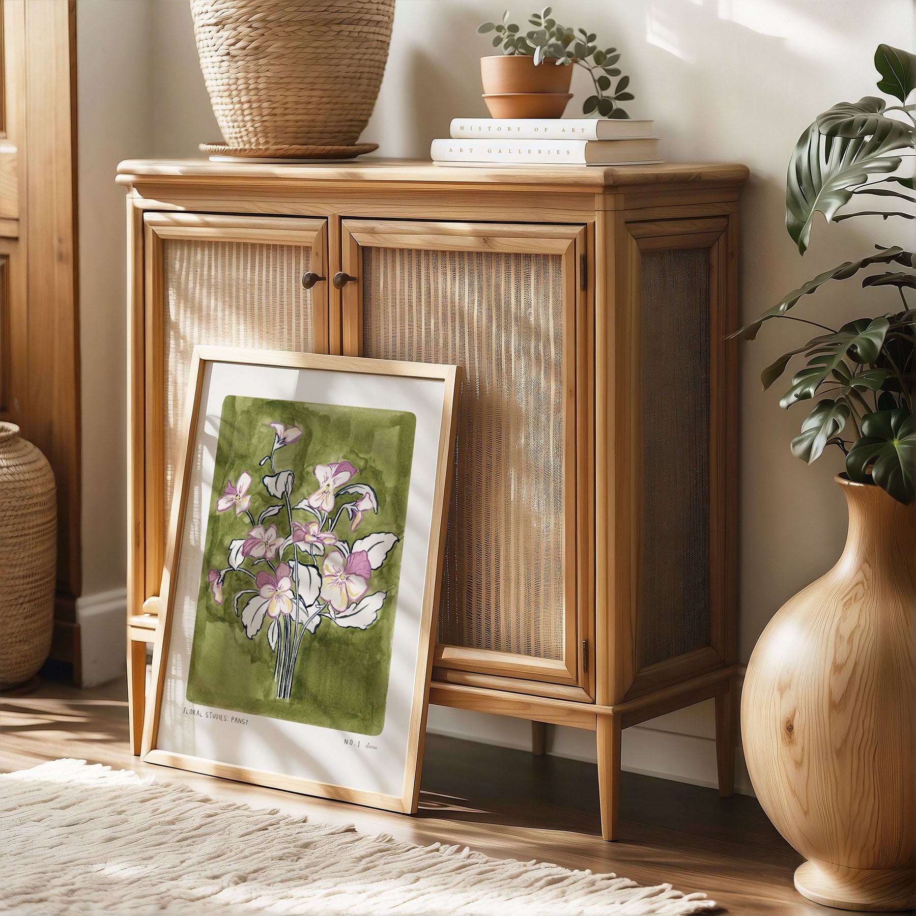 Pansy Art Print, a olive green painted background with light purple and light yellow pansy flower heads with navy outlined leaves. With small text reading Floral studies: pansy NO.1 styled in a light wood frame against rattan furniture