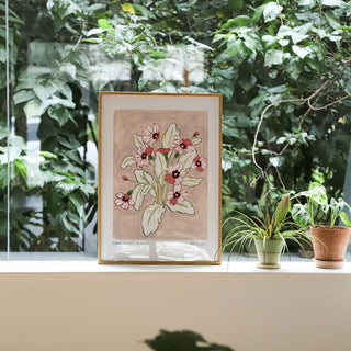 Primrose art print, a dusky light pink painted background with rosey pink and deep maroon flowers and olive green leaves. With small text reading floral studies:primrose NO.4 styled in a light wood frame against plants and greenery