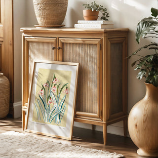 Snowdrop art print, a light muted yellow painted background with pinky snowdrop flowers and olive green/ turquoise leaves. With small text reading floral studies: snowdrop no.2 styled in a light oak frame against rattan furniture