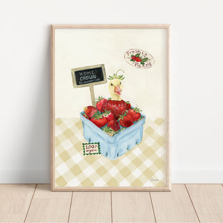 A framed watercolor art print leaning against a white wall on a light wood floor. The illustration shows a blue crate of red strawberries on a beige-and-white checkered table, with a small duck and signs reading 'Home grown Organic' and 'Freshly Picked'.