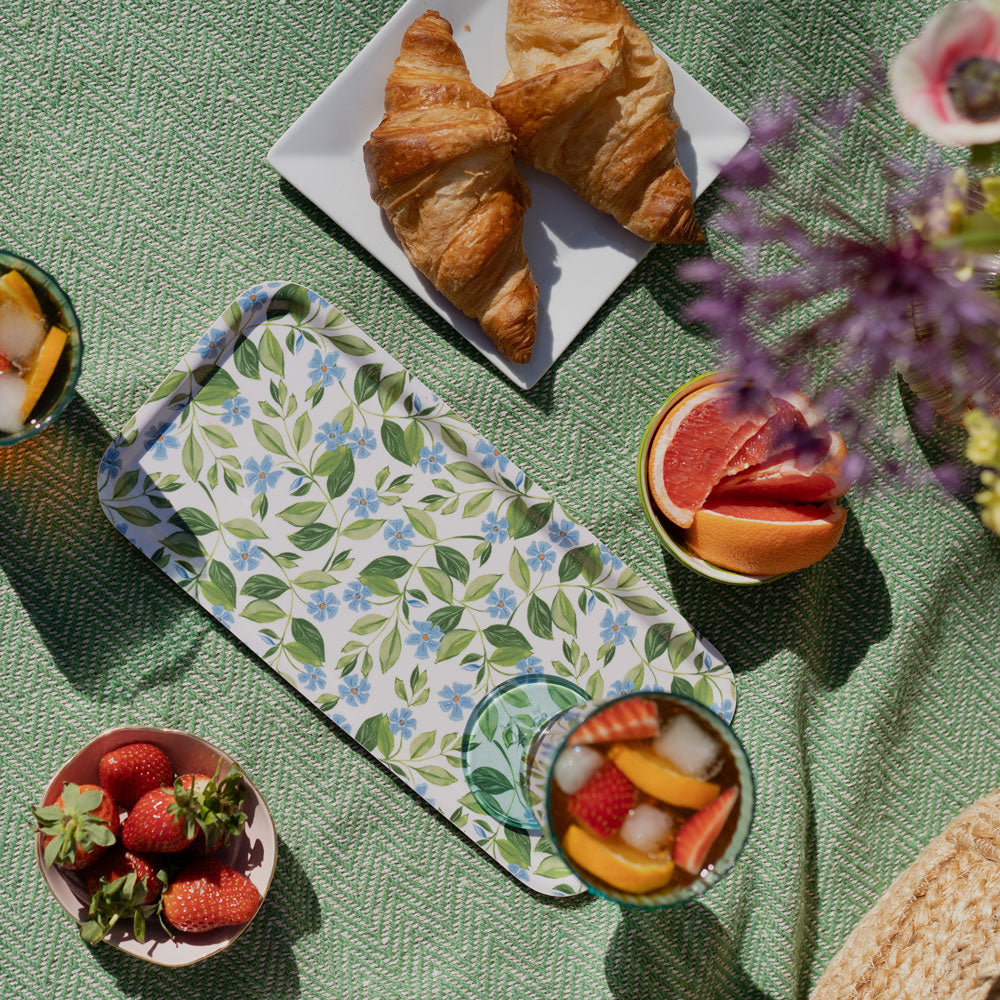 picnic flatlay, tray holding fruit filled drink. photographed on a green rug with jug of flowers and picnic foods scattered around