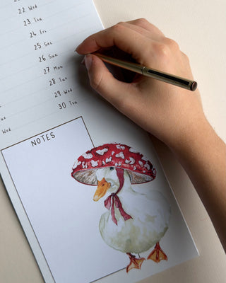A hand holding a gold pen writes on a duck wall calendar, with a printed page below featuring a watercolor illustration of a white duck wearing a red mushroom bonnet 