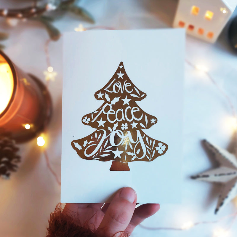 gold foil christmas tree with text love peace joy cut out in white. paired with a brown envelope.